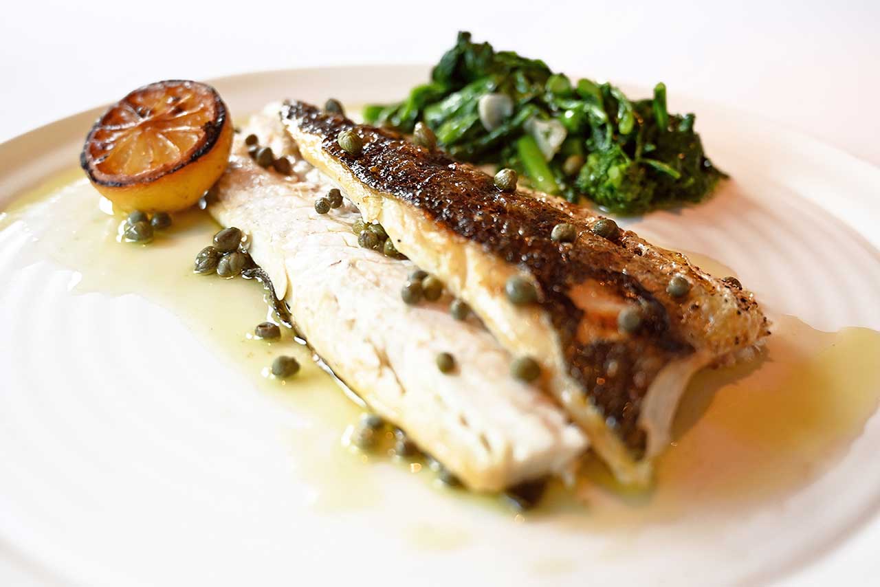 grilled fish on a whit plate with capers, greens and lemon